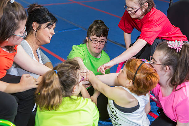 Group of special needs teenagers and young women with instructor, showing team spirit. They are sitting in a circle on a gym floor, hands in the center. Several of the girls have downs syndrome and two are in wheelchairs.